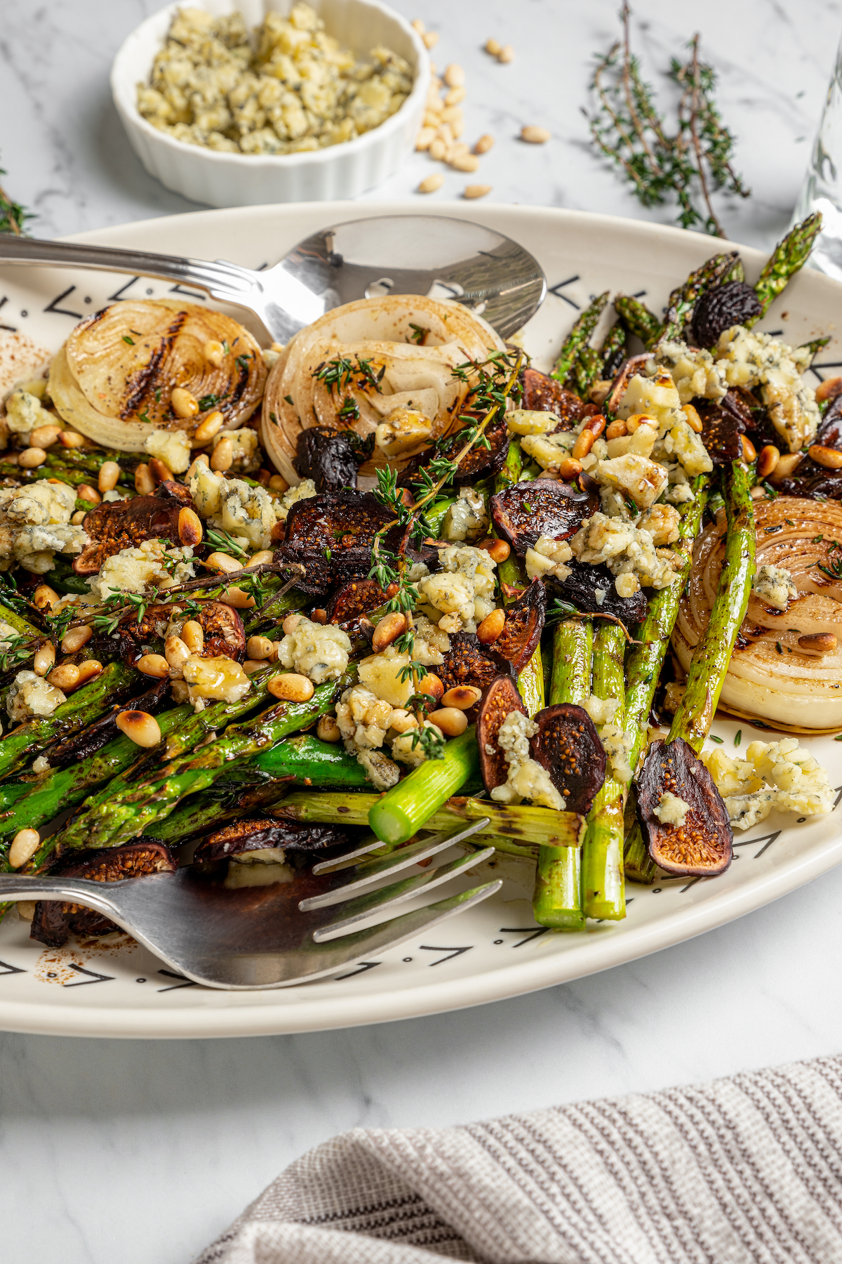 Grilled Asparagus with Onion recipe is topped with simple vinaigrette, blue cheese, black Mission figs, and toasted pine nuts. So flavorful! A homemade grilled asparagus salad on an oval platter.