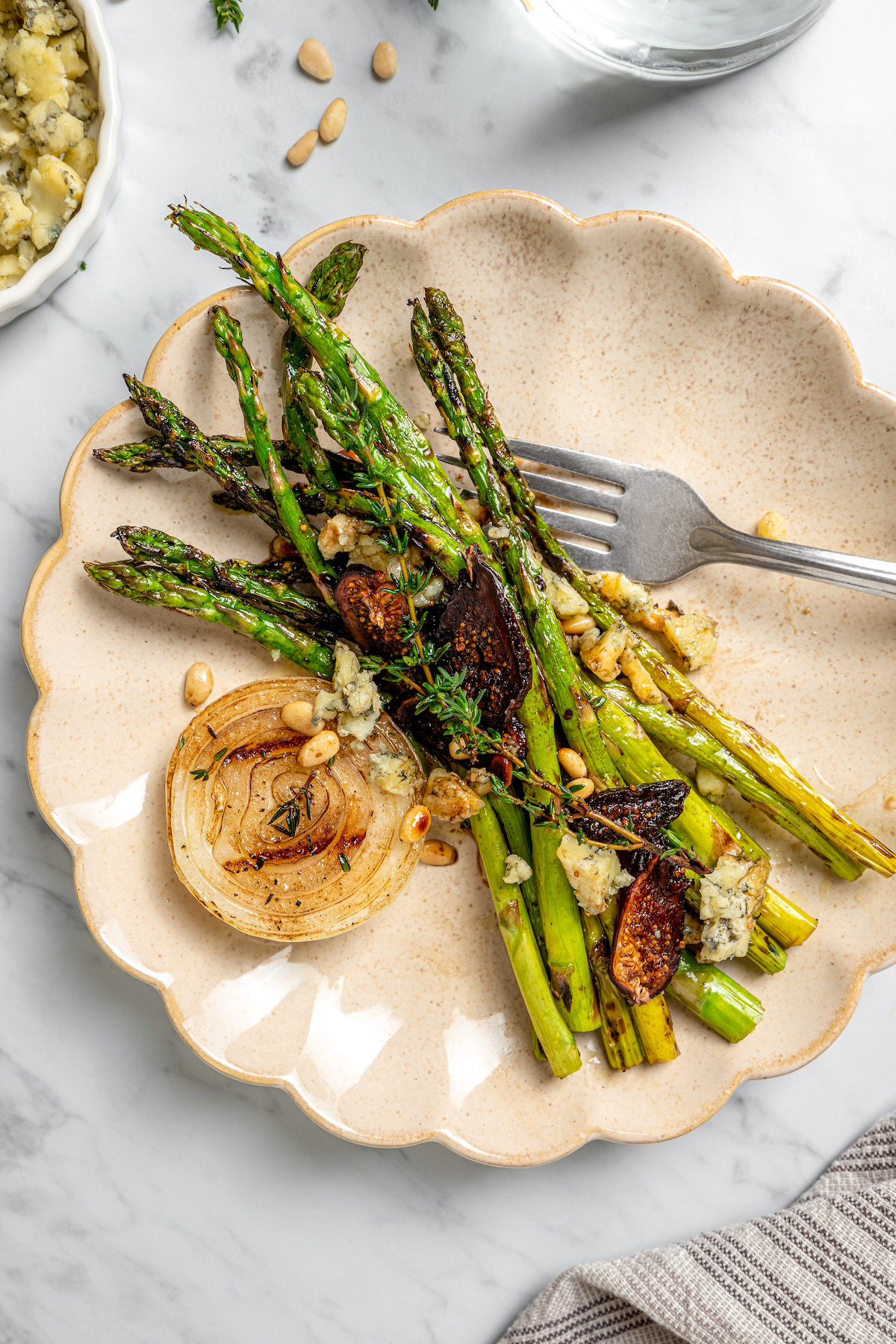 Grilled Asparagus with Onion recipe is topped with simple vinaigrette, blue cheese, black Mission figs, and toasted pine nuts. So flavorful! A serving of asparagus salad on a plate with scalloped edges.