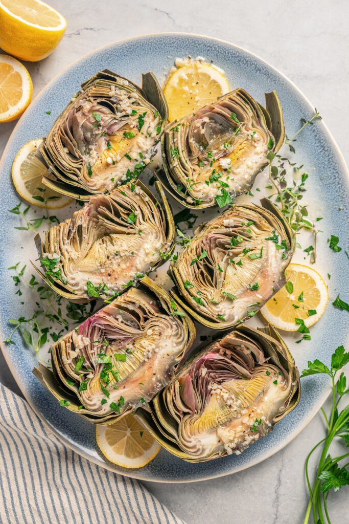 This tender Instant Pot artichokes recipe with hints of tangy lemon, garlic, and fresh herbs will be your go-to holiday side dish. Halved artichokes with garlic, fresh herbs, and lemon wedges.